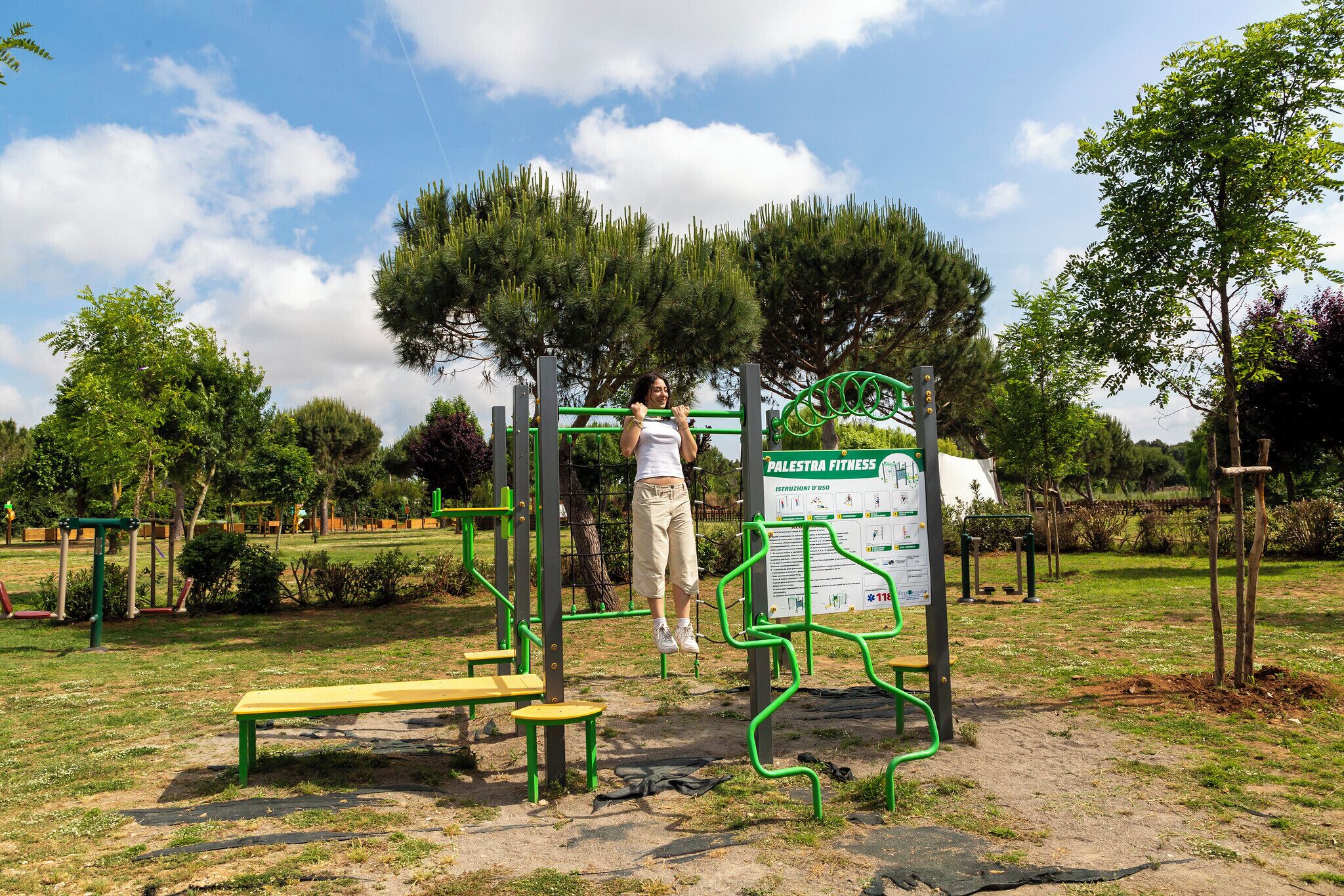 Un parque con equipo de ejercicios, un banco verde y un cartel de Palestra Fitness.