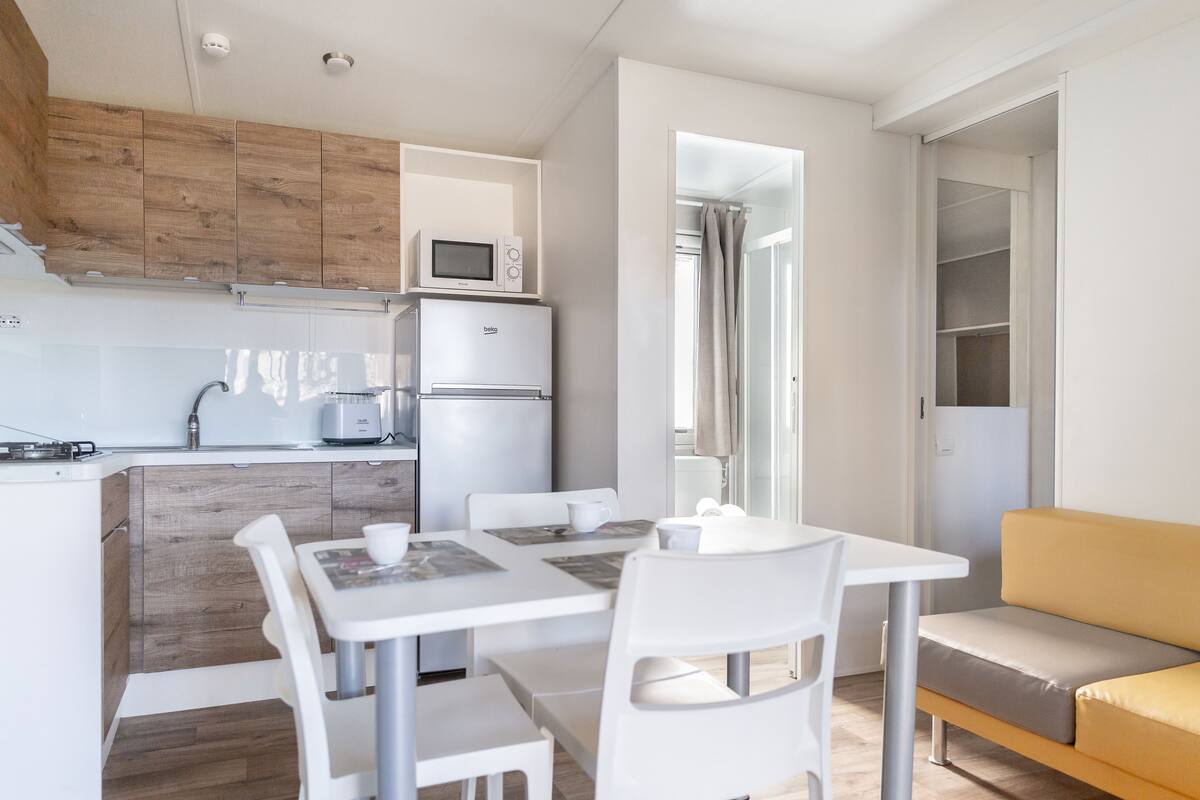 Cocina compacta con microondas, refrigerador y gabinetes de madera. Hay un comedor con mesa y sillas, y un sofá en la sala.