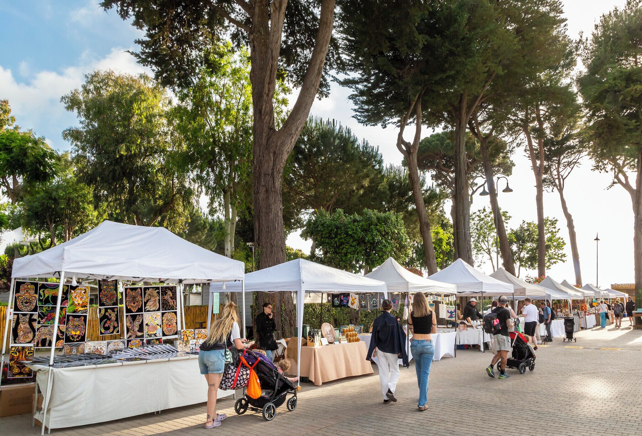 A row of white tents with tables displaying artwork, set up in an outdoor market area with trees in the background.