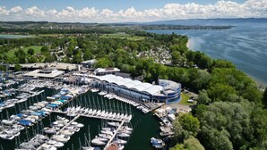 Un port de plaisance où sont amarrés de nombreux bateaux, un grand bâtiment doté d’un toit incurvé, et une vue sur un lac et la végétation environnante.