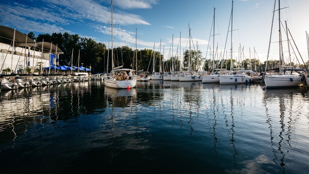 Un port de plaisance où sont amarrés de nombreux voiliers, un bâtiment doté d’une marquise bleue et une eau calme qui reflète les bateaux.