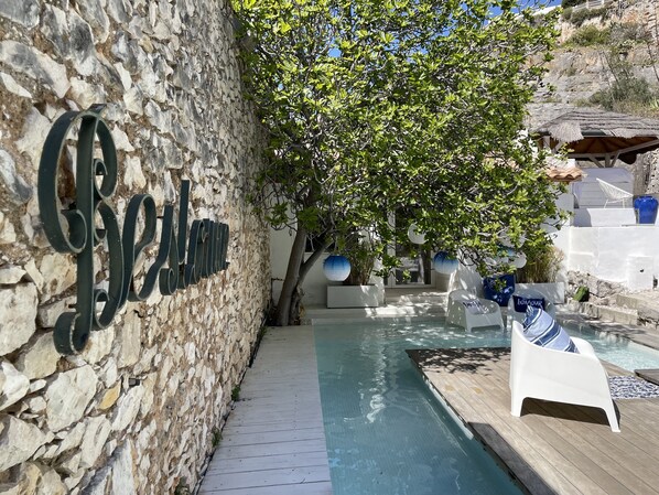 Un mur de pierre avec une pancarte, un espace piscine avec des chaises longues et un arbre qui offre de l’ombre.