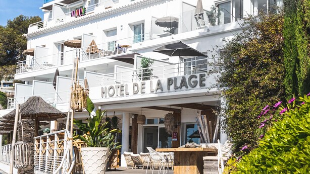 Un hôtel doté de nombreux balcons, d’un espace extérieur aménagé pour s’asseoir et d’une enseigne indiquant « HÔTEL DE LA PLAGE ».