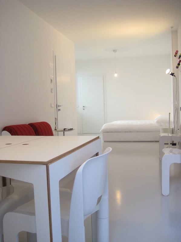 A modern, minimalist bedroom with a white bed, a desk, and a chair.