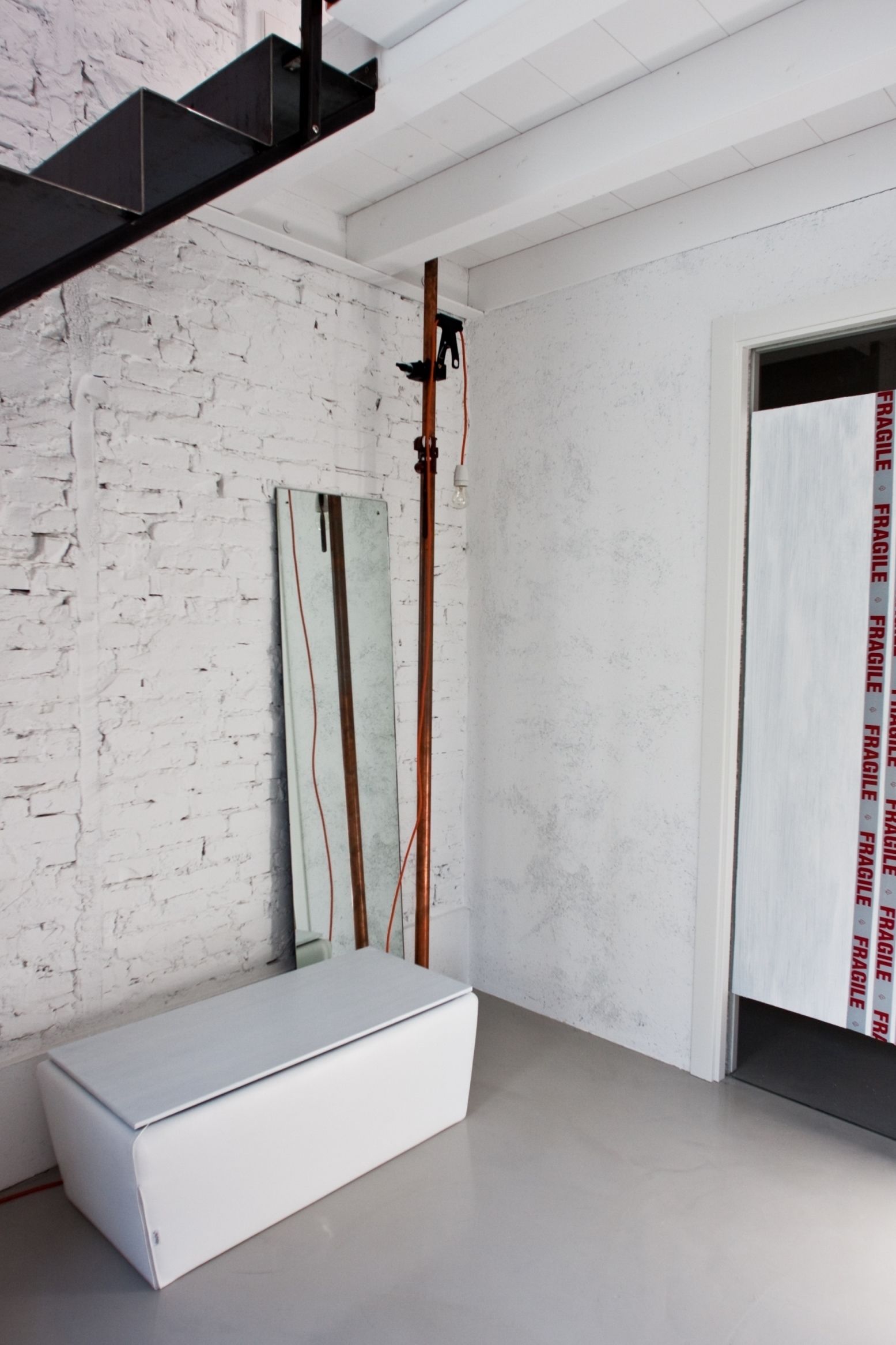 Une pièce avec un mur en briques blanches, un banc blanc et une porte sur laquelle est apposée une étiquette indiquant « FRAGILE ».