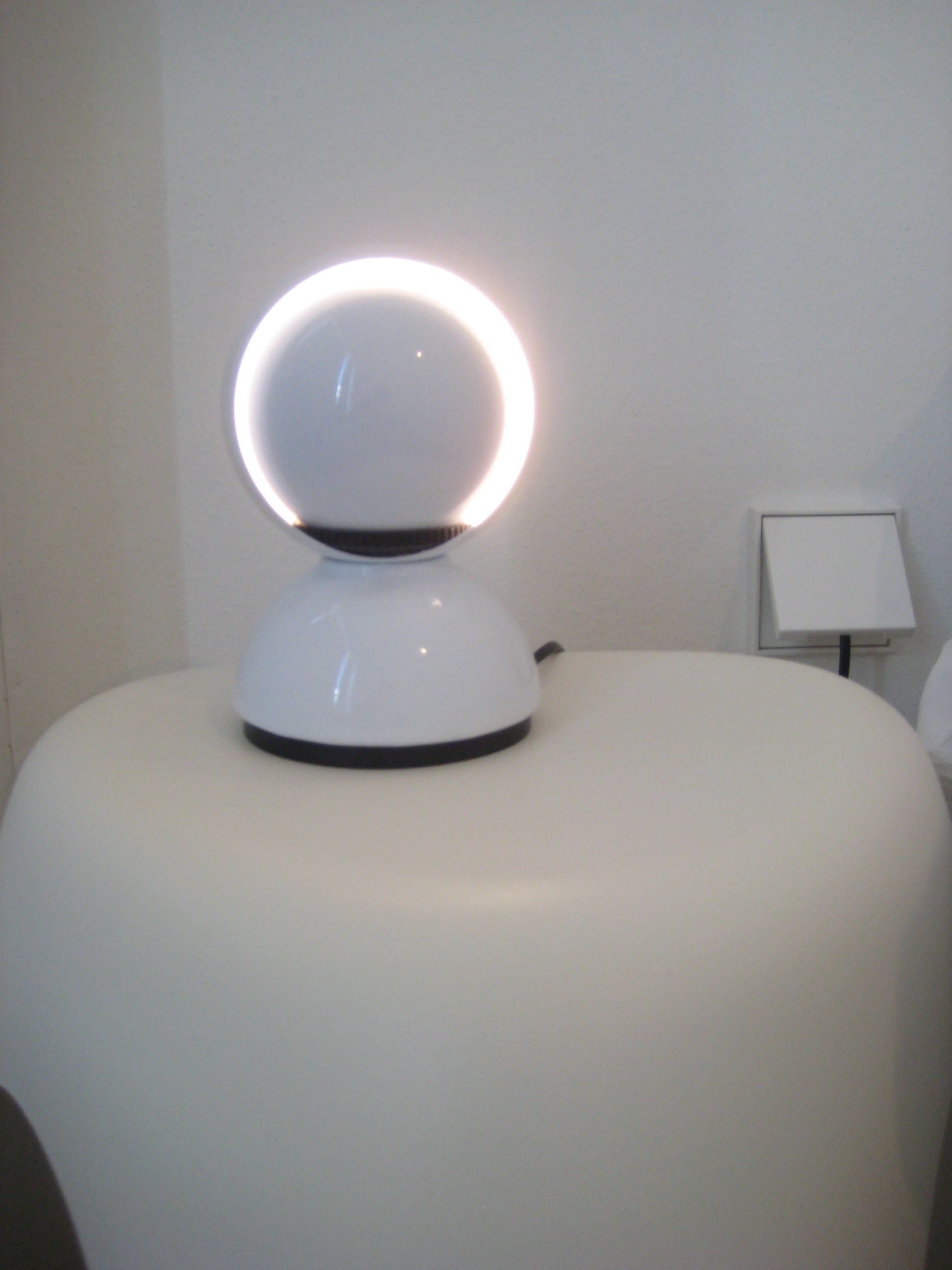 Un objet blanc, rond et lumineux, posé sur une surface blanche, qui pourrait être une lampe, avec un petit objet blanc et rectangulaire à sa droite.