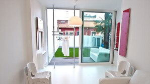 A modern room with white furniture, a sliding glass door to a balcony, and a view of a house with a red roof. - Legrenzi Rooms (Mestre)