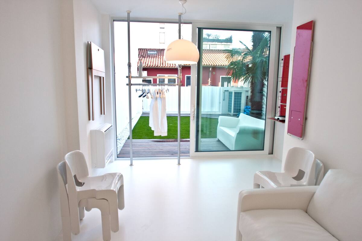Ein modernes Zimmer mit weißen Möbeln, einer Schiebetür zum Balkon und Blick auf ein Haus mit rotem Dach.
