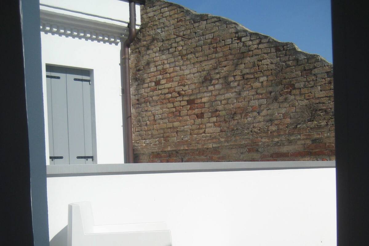 Eine Holzterrasse mit einem weißen Stuhl und einem weißen Tisch. Im Hintergrund ist eine Ziegelwand zu sehen.