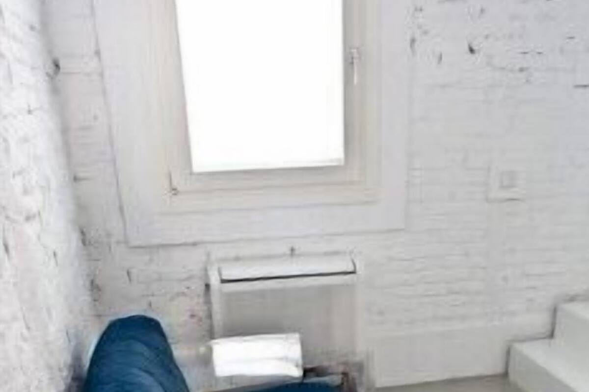 Ein Zimmer mit weißer Backsteinwand, einem Fenster, einem blauen Sofa, einem kleinen runden Tisch mit einem roten Gegenstand und einem weißen Heizkörper.