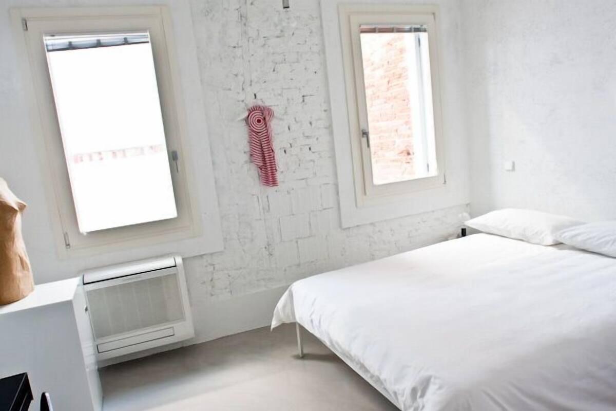 Ein Schlafzimmer mit weißer Ziegelwand, zwei Fenstern, einem Bett und einer Heizung.
