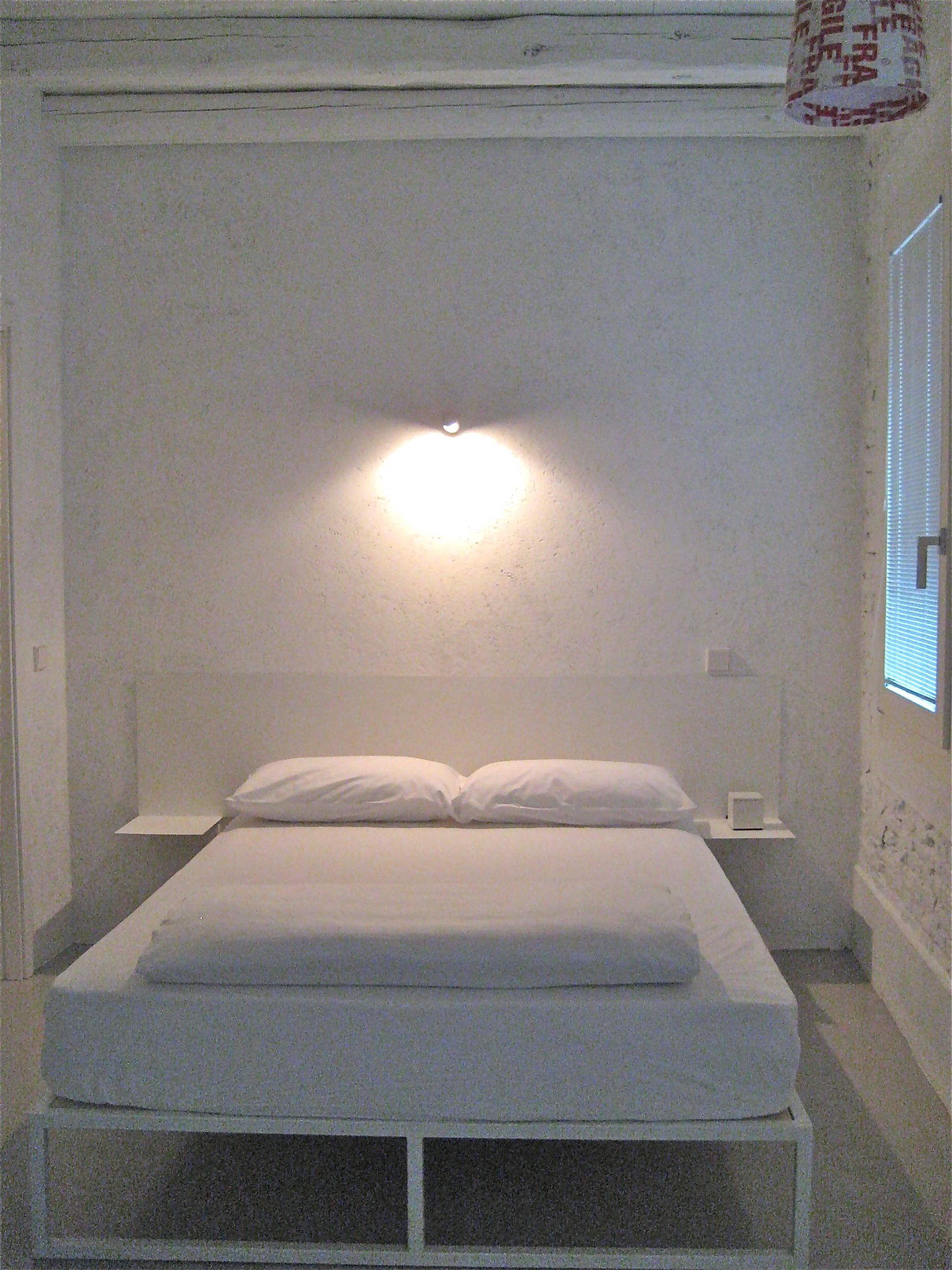 Ein kleines, weißes Zimmer mit einem Einzelbett, schlichtem Design, einem Nachttisch und einer Wandleuchte.