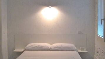 Ein kleines, weißes Zimmer mit einem Einzelbett, schlichtem Design, einem Nachttisch und einer Wandleuchte.