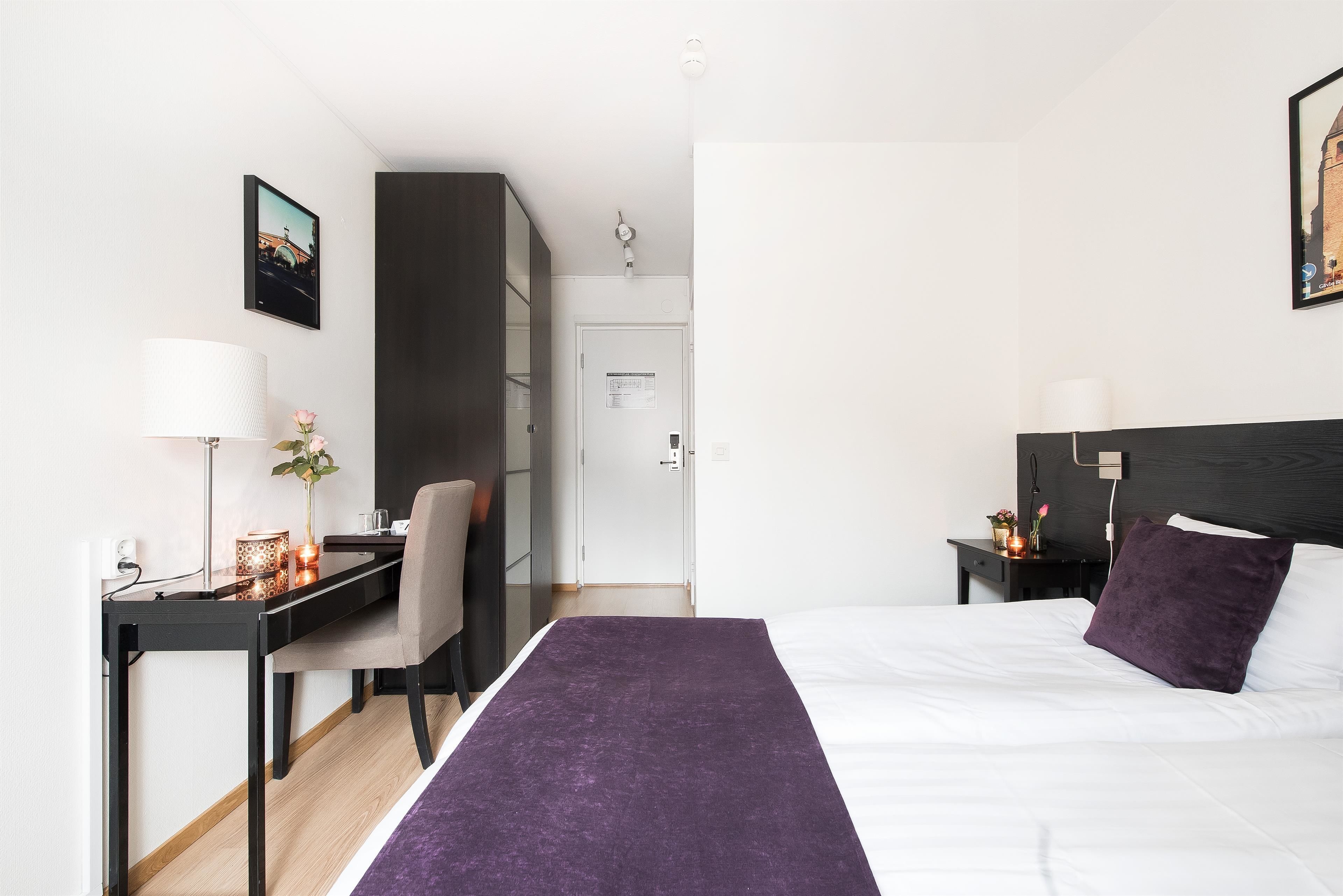 A modern hotel room with a bed, a desk, a chair, a wardrobe, and a nightstand with a lamp.