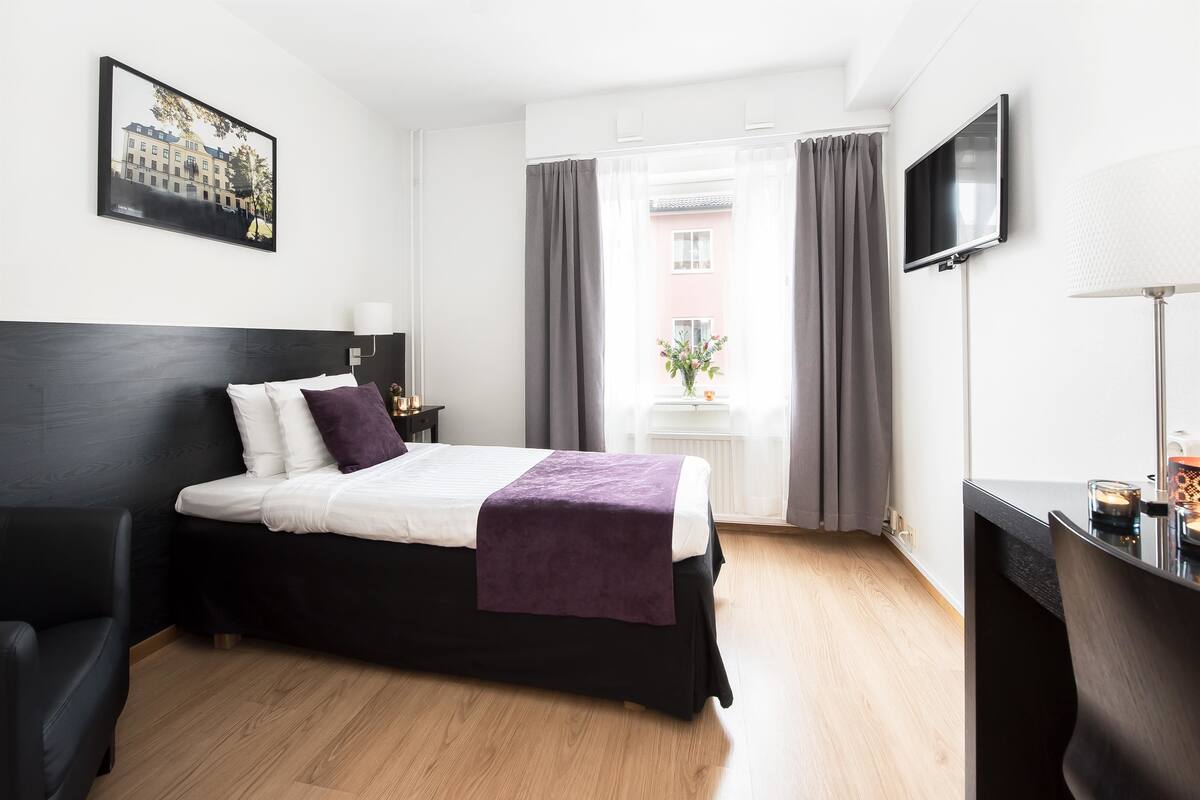Habitación de hotel con cama, escritorio, televisor y ventana con cortinas.