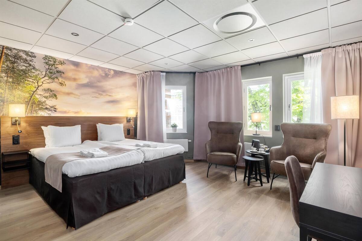 Habitación de hotel con una cama grande, dos sillones, una mesita y vistas a árboles a través de las ventanas.