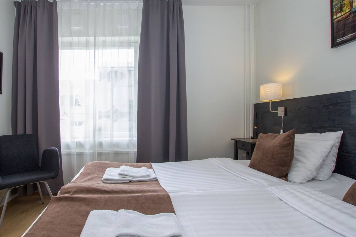 Una habitación de hotel con cama, una silla, televisión, lámpara y ventana con cortinas.