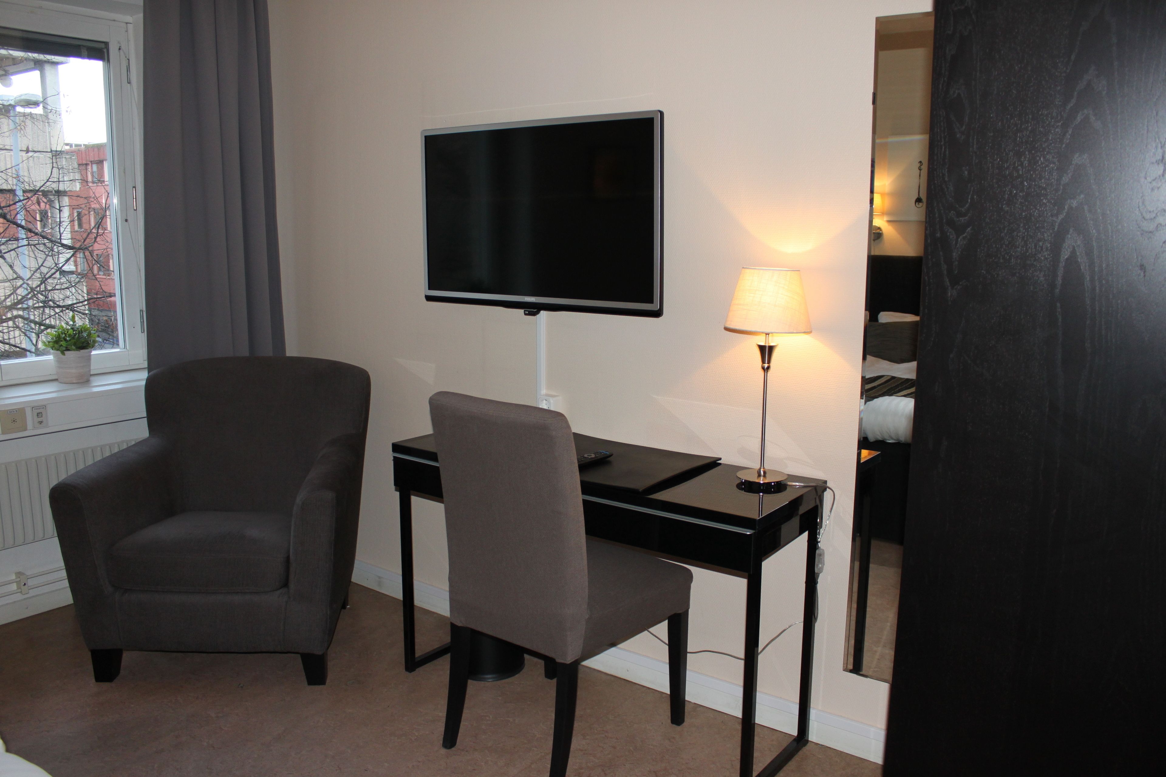 A hotel room with a desk, a chair, a TV, a lamp, and a window with a view of buildings.
