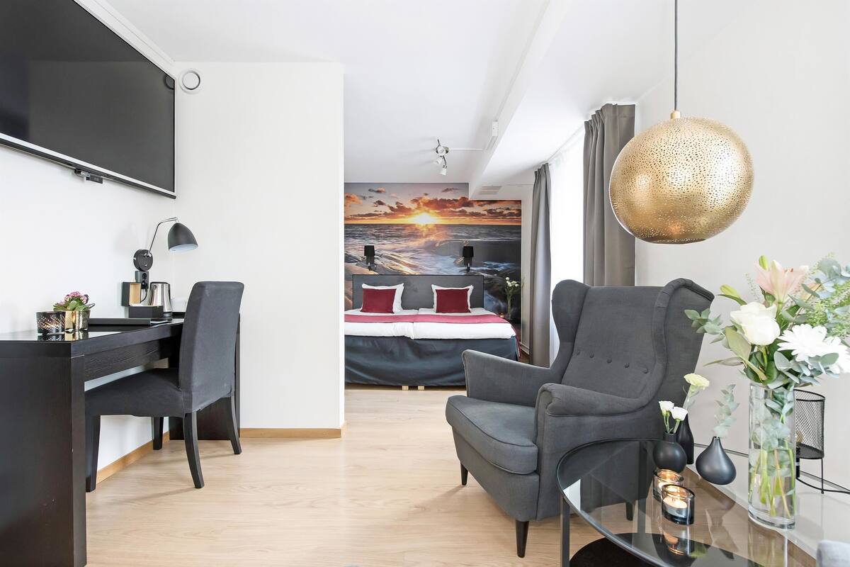 Habitación de hotel moderna con una televisión de pantalla plana grande, un escritorio con silla, un sillón gris, una mesa de centro de vidrio y un jarrón decorativo con flores.