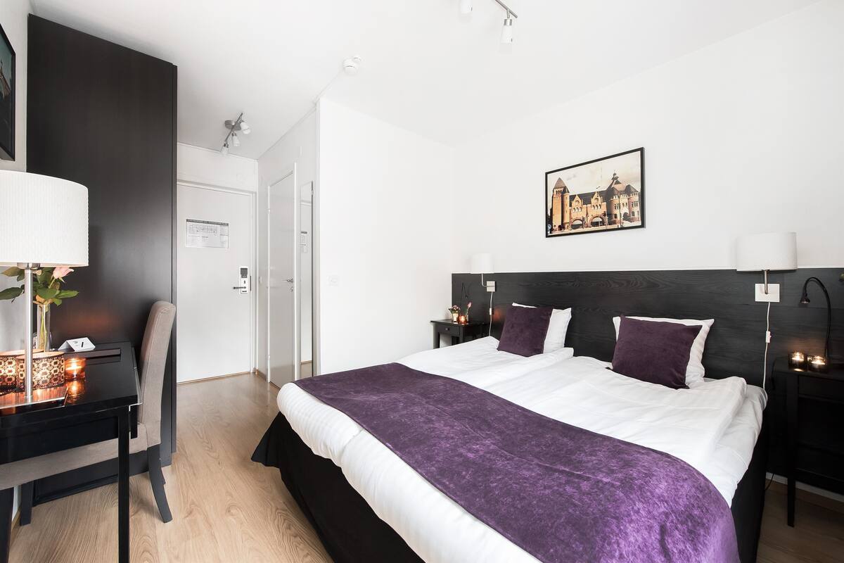 Una habitación de hotel moderna con una cama grande, un escritorio con lámpara y una mesita de noche con lámpara.