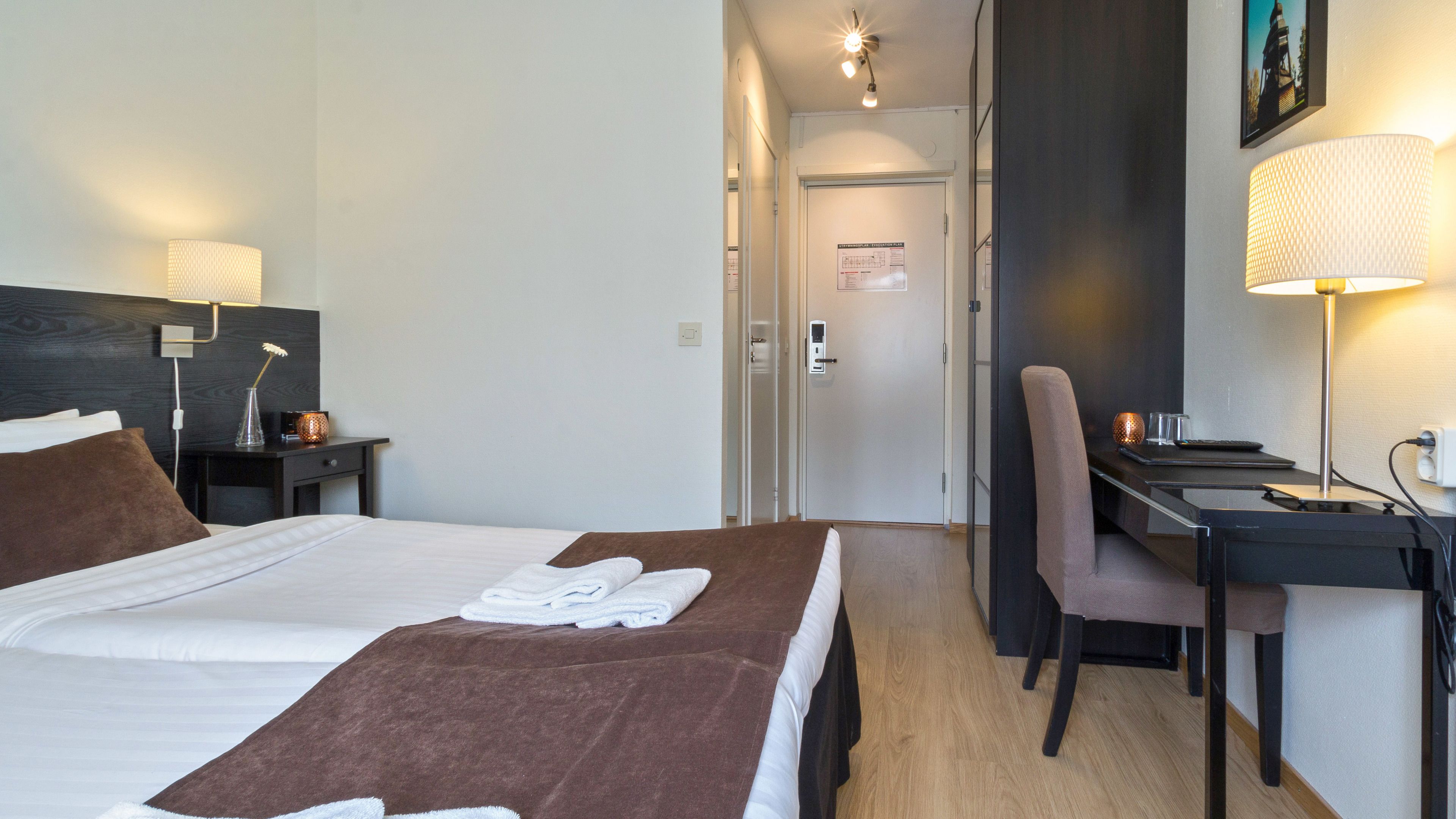 Ein Hotelzimmer mit einem Bett, einem Schreibtisch und einem Stuhl. Auf dem Schreibtisch steht eine Lampe und auf dem Nachttisch ebenfalls.