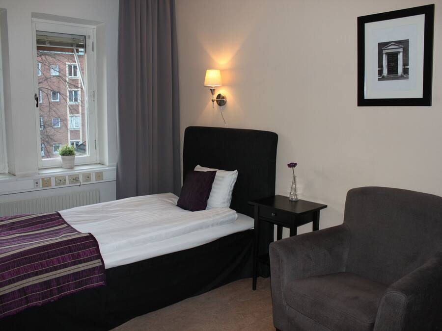 Ein Hotelzimmer mit Bett, Sessel, Nachttisch, Lampe und einem gerahmten Bild an der Wand.