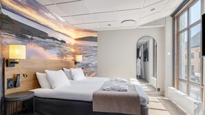 Een hotelkamer met een groot bed, uitzicht op de stad door grote ramen, een muurschildering van een zonsondergang boven het water, en een klein nachtkastje met een lamp.
