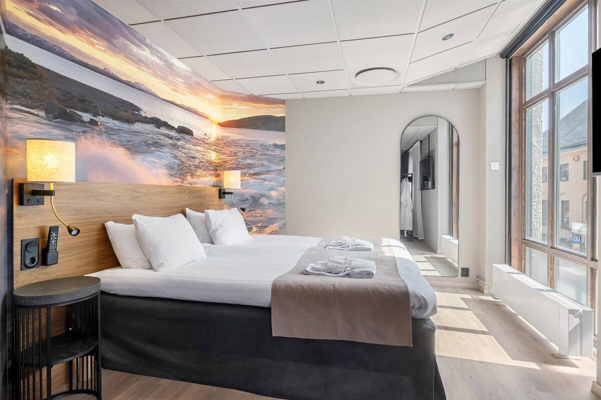 Habitación de hotel con una cama grande, vistas de la ciudad a través de amplias ventanas, un mural de un atardecer sobre el agua en la pared y una mesita de noche con lámpara.