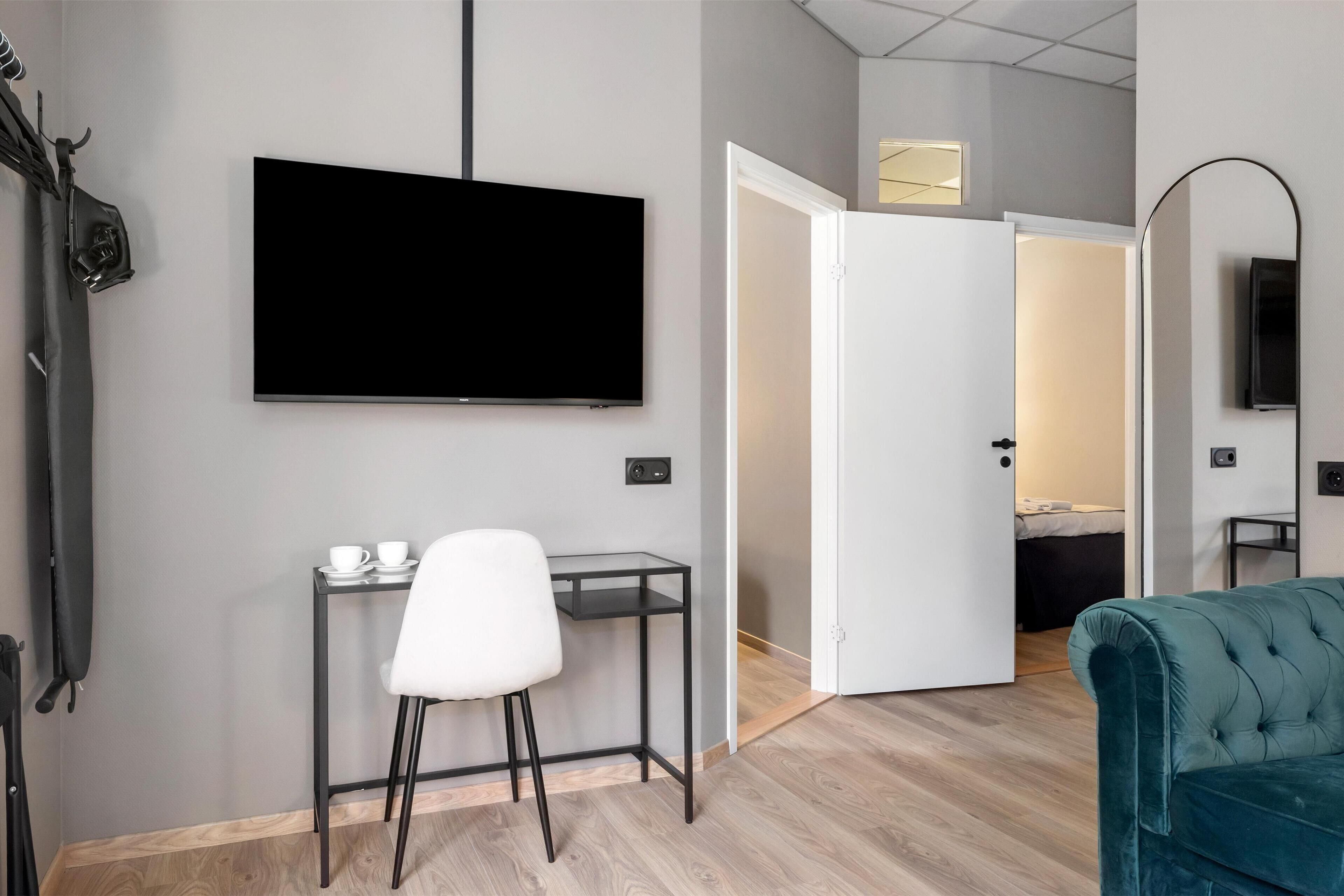 Une chambre d’hôtel moderne équipée d’un téléviseur à écran plat fixé au mur, d’un petit bureau avec une chaise, d’un canapé de couleur turquoise et d’une porte donnant sur une autre pièce.