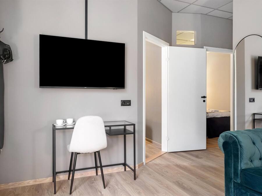 A modern hotel room with a flat-screen TV mounted on the wall, a small desk with a chair, a teal sofa, and a door leading to another room.