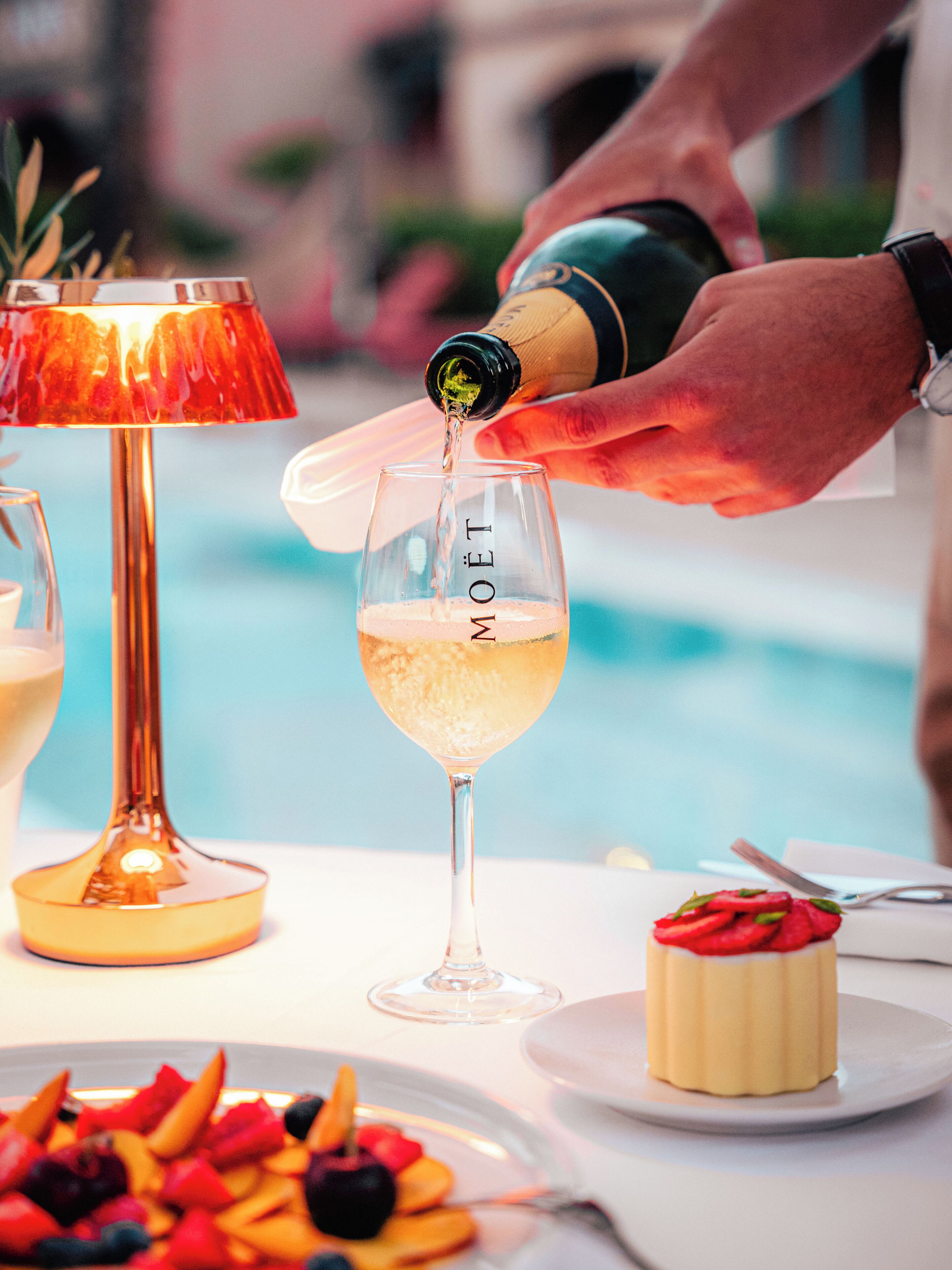 Una persona versa champagne in un bicchiere con l'etichetta "MOËT" a un tavolino a bordo piscina, con frutta e dessert.