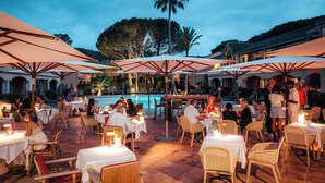 Un espacio de comedor al aire libre con mesas y sillas, sombrillas y una piscina al fondo.