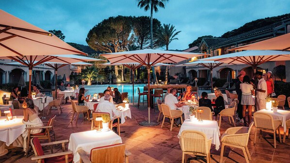 Un espacio de comedor al aire libre con mesas y sillas, sombrillas y una piscina al fondo.