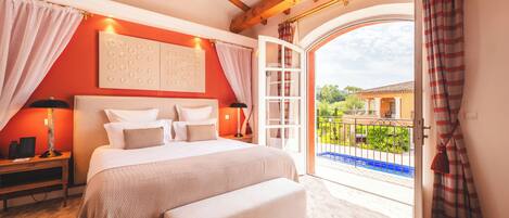A bedroom with a large bed, a bench, a nightstand, a wall-mounted light, and a view of a balcony with a railing.