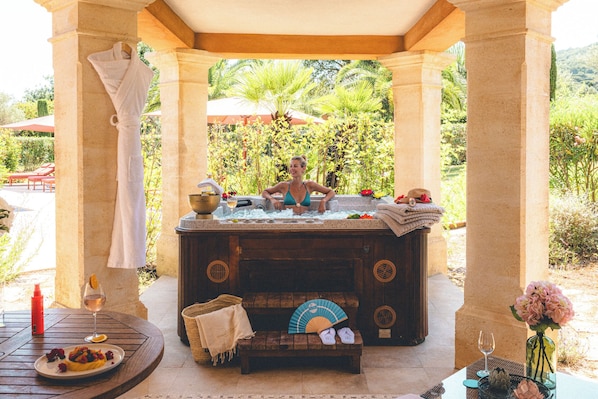 Estructura de piedra con un jacuzzi, una mesa con un plato de comida y un banco con un ventilador y zapatillas.