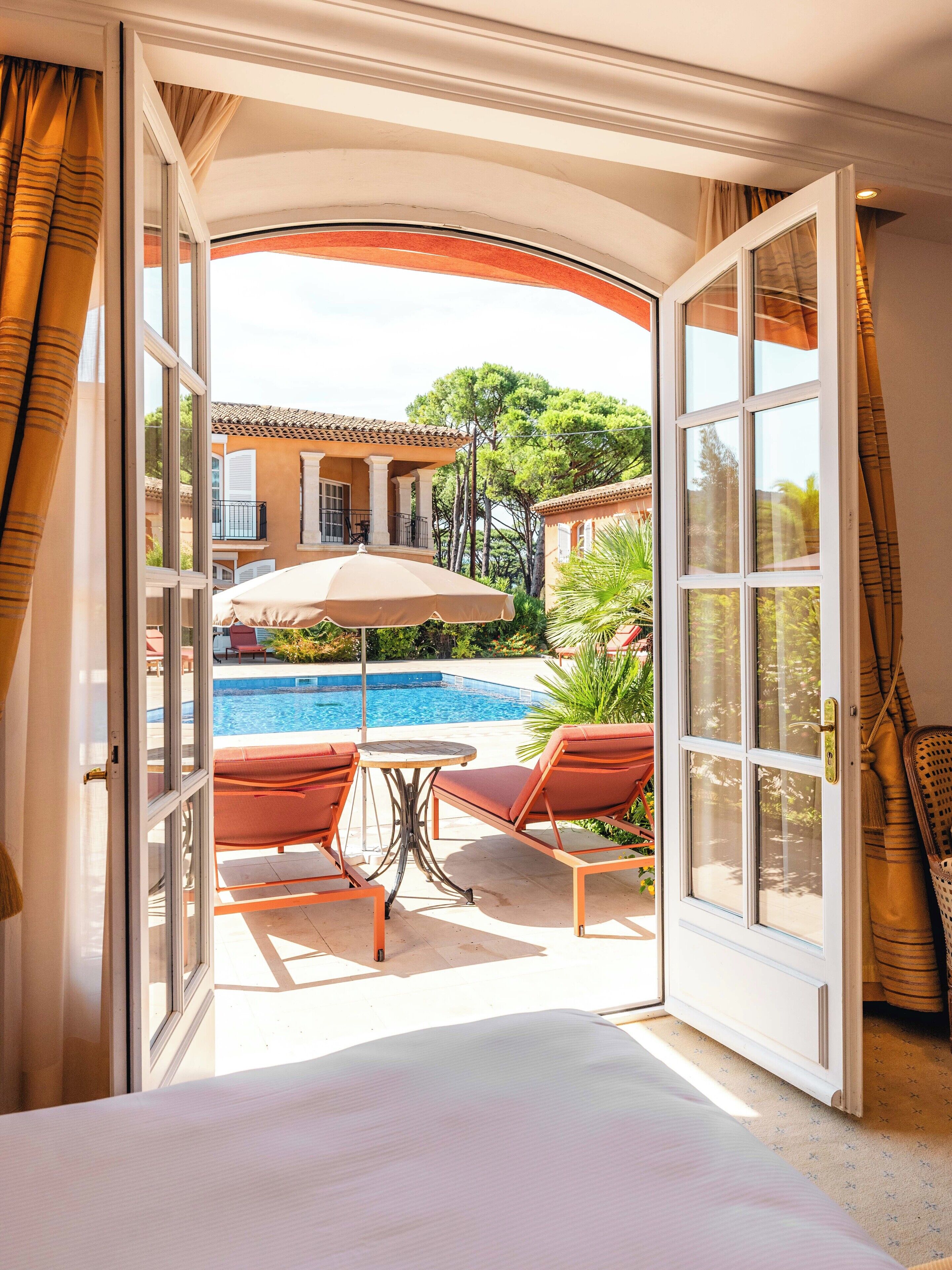 A bedroom with an open door leading to a pool area, featuring a bed, two lounge chairs, and an umbrella.