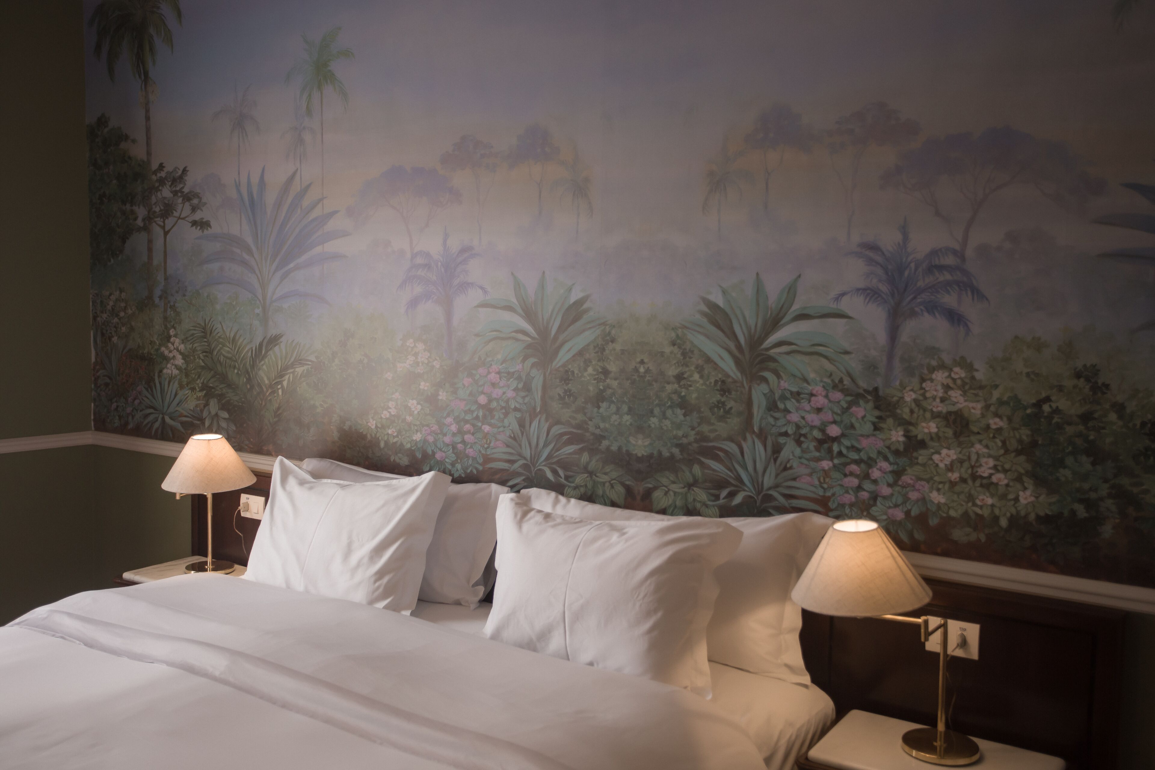 Una habitación de hotel con una cama, dos lámparas de noche y un mural de paisaje tropical en la pared.