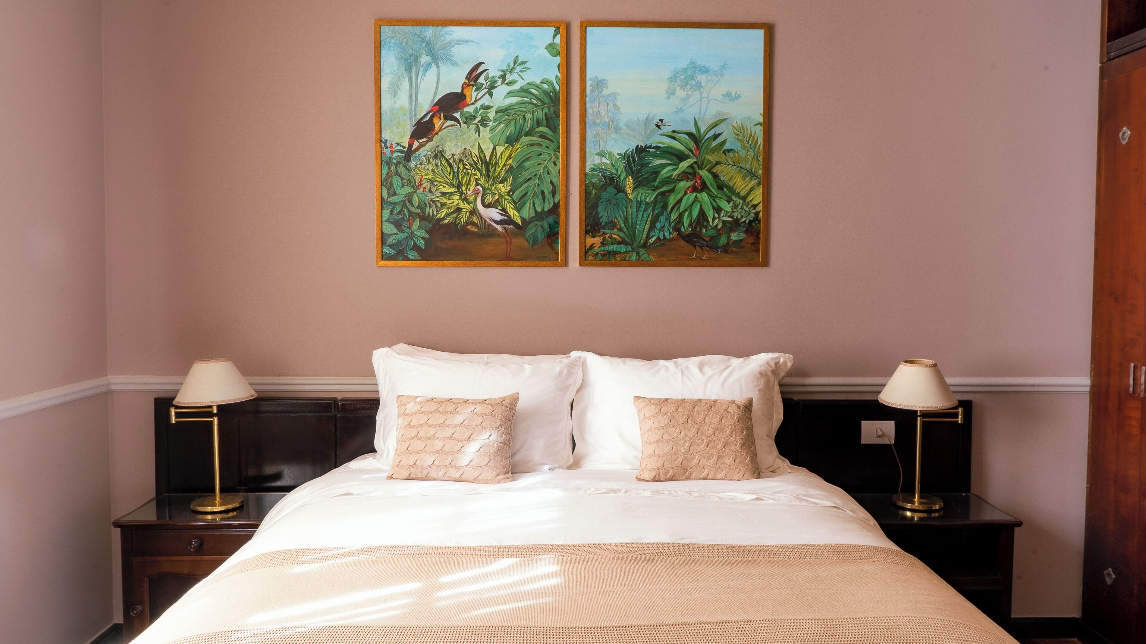 Una habitación de hotel con una cama, dos mesitas de noche, dos lámparas y dos cuadros con aves tropicales en la pared.