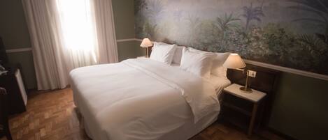 A hotel room with a large bed, two bedside lamps, a nightstand, a wooden floor, a window with curtains, and a mural on the wall.