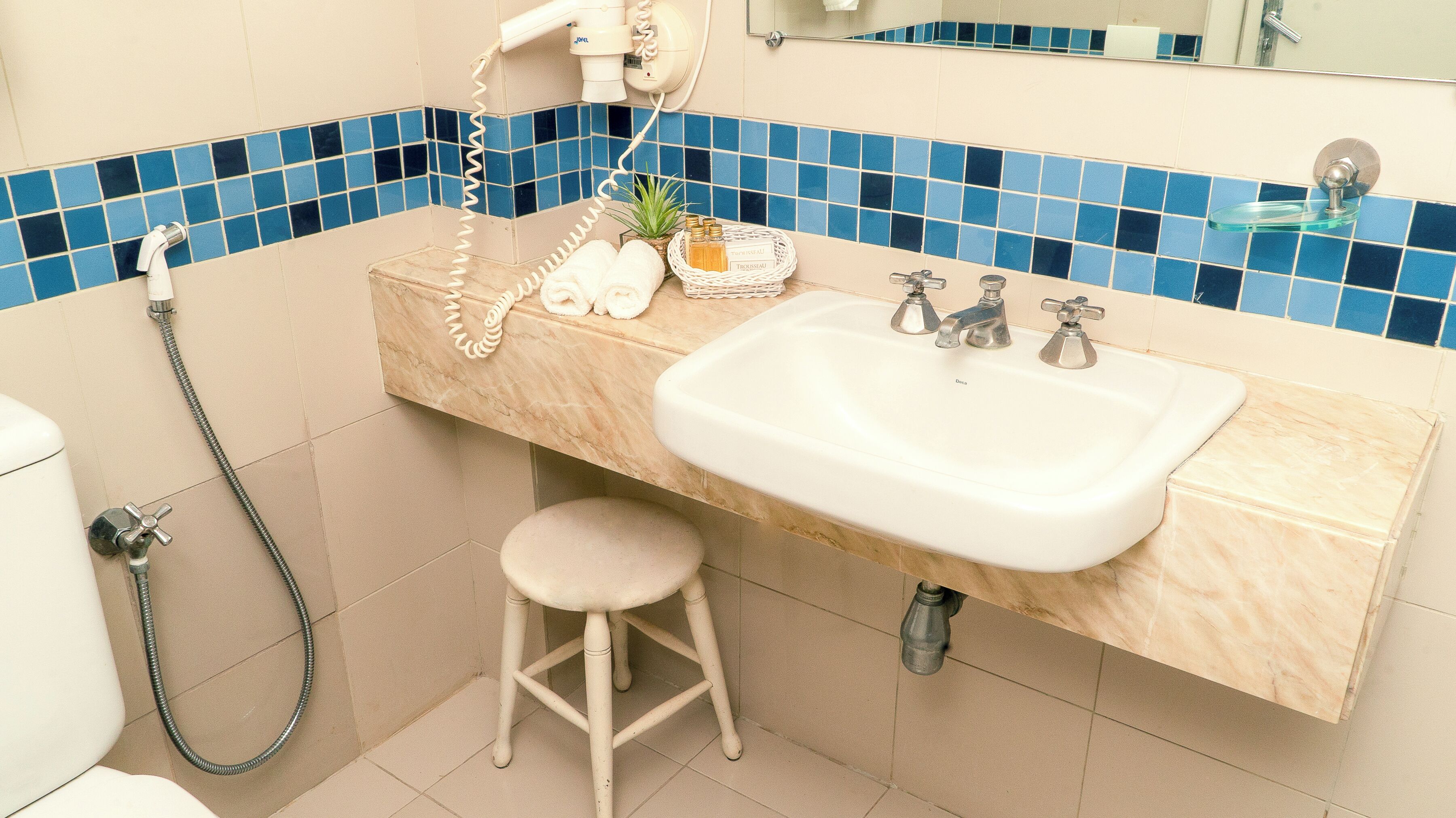 Un baño con lavabo, dos grifos, un secador de pelo y un taburete pequeño.
