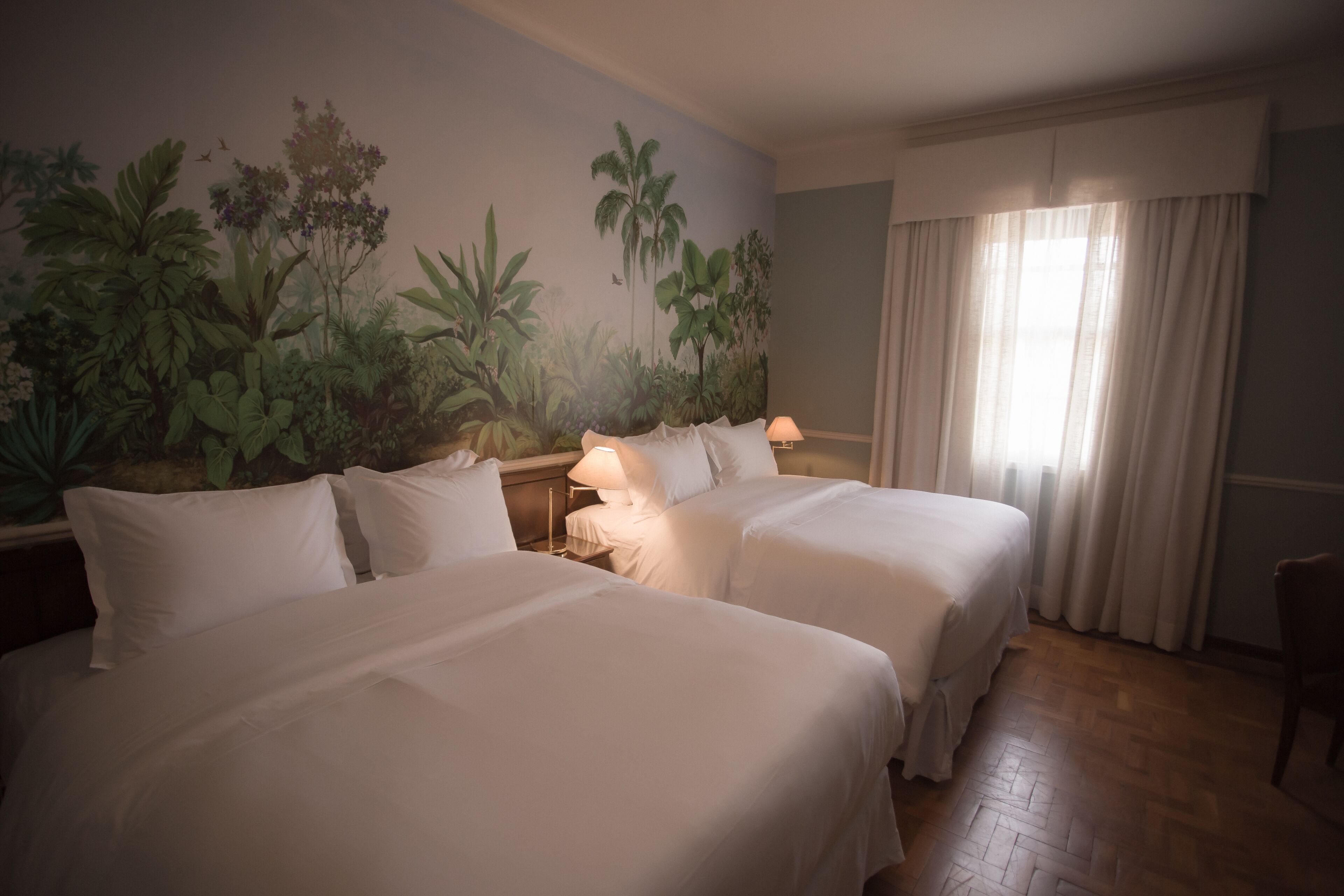 A hotel room with two beds, a nightstand, a lamp, a window with curtains, and a wall mural of tropical plants.