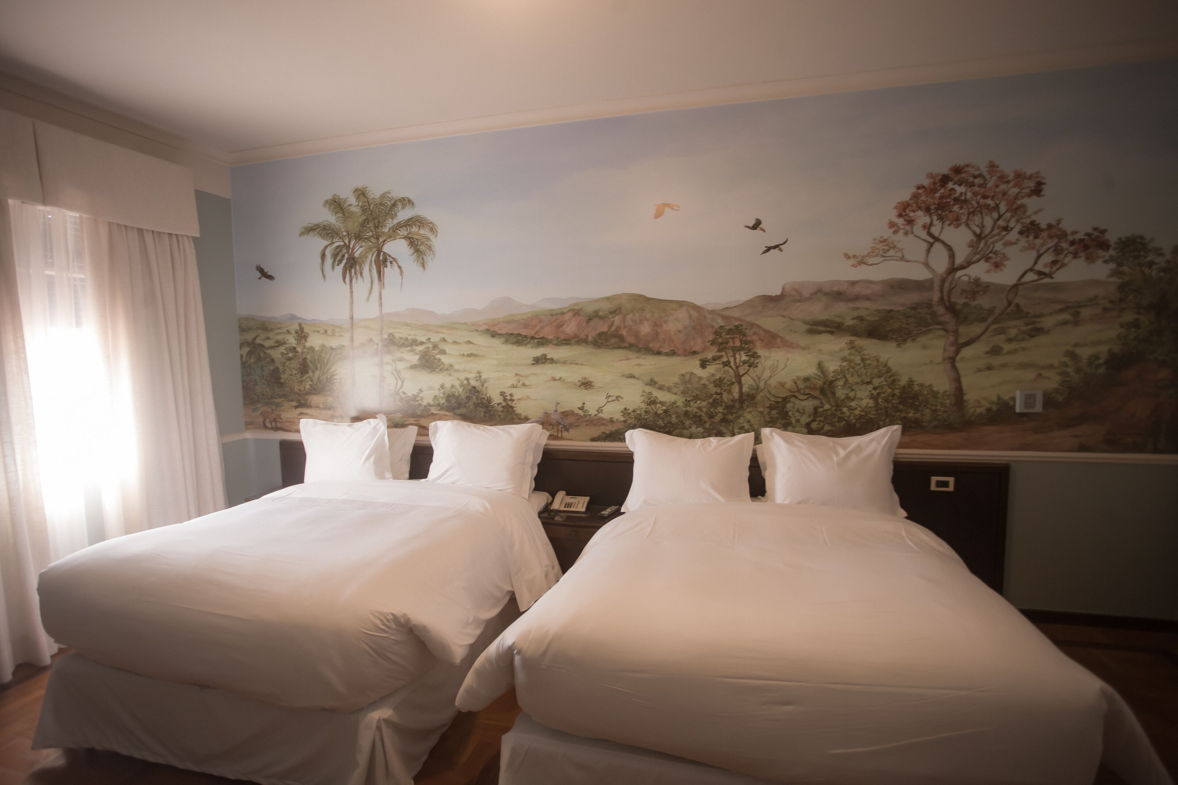 Een hotelkamer met twee enkele bedden, een muurschildering van een landschap en een raam met gordijnen.