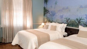 Ett hotellrum med två sängar, ett stort fönster med tunna gardiner, en väggmålning med tropiskt landskap och en sänglampa.
