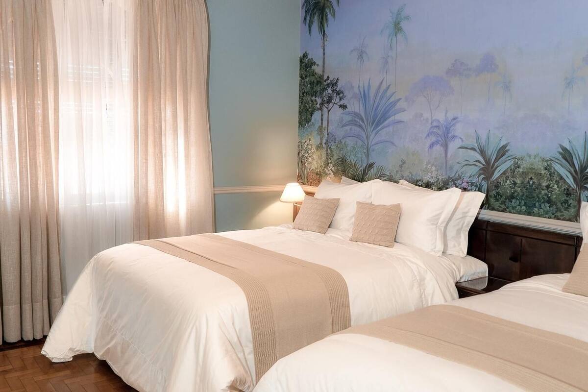Quarto de hotel com duas camas, uma janela grande com cortinas leves, um mural na parede com paisagem tropical e uma luminária de cabeceira.