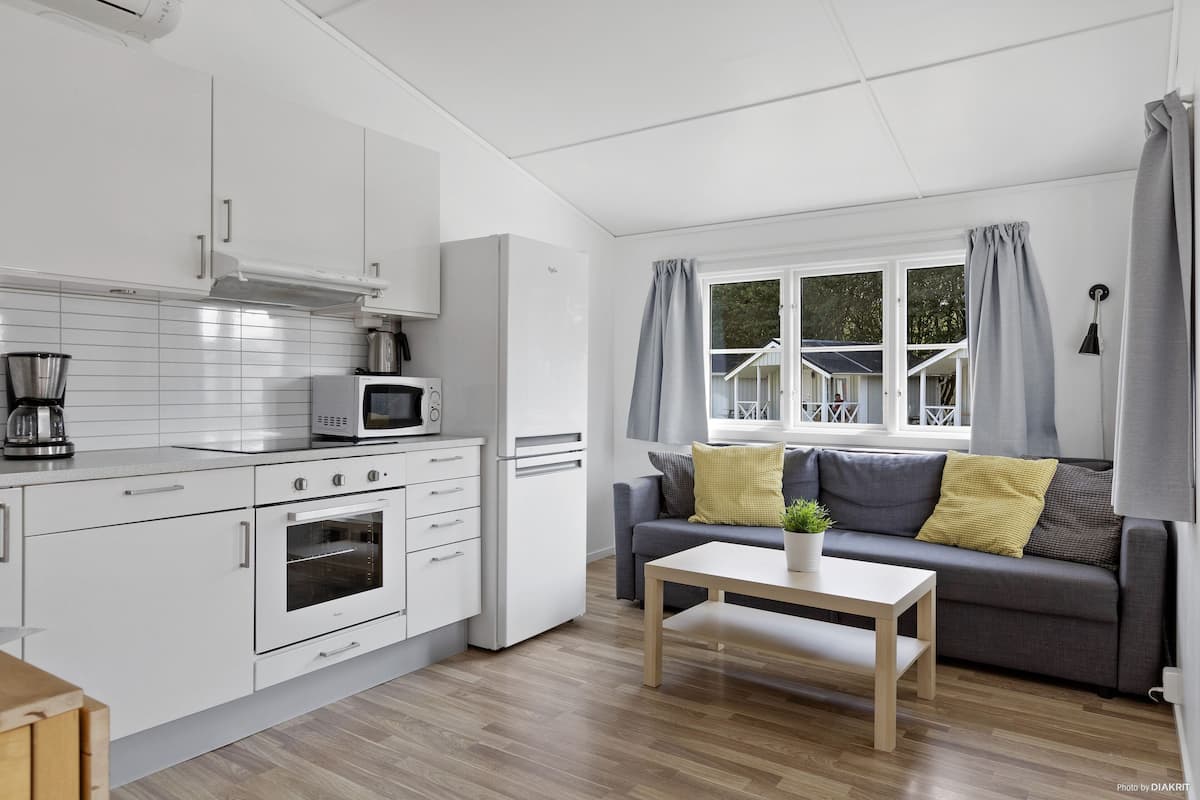 A modern kitchen with white cabinetry, a microwave, and an oven. There is a gray sofa with yellow pillows and a small table with a plant.
