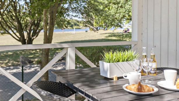 A wooden deck with a table set for breakfast, including croissants, coffee cups, and wine glasses, overlooking a grassy area with trees and a body of water. - First Camp Malmö (Malmö)