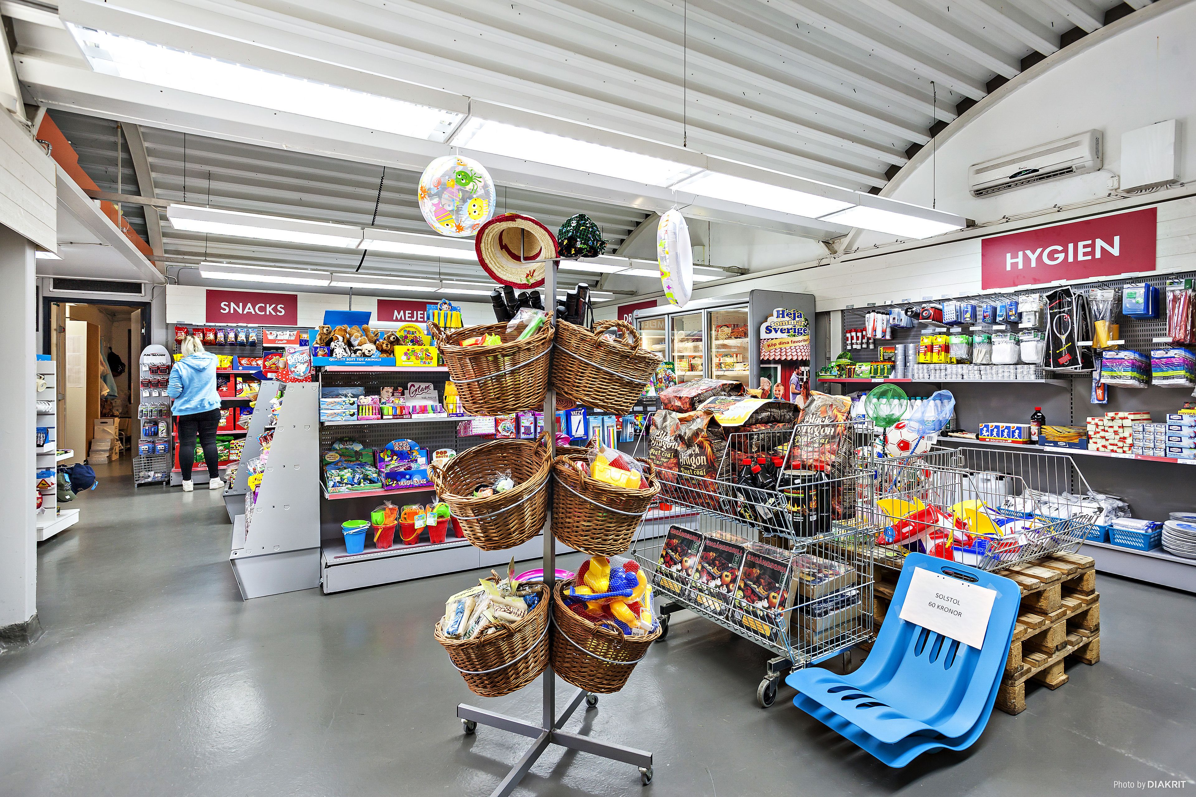 Een winkel met gangpaden met de aanduiding 'SNACKS' en 'HYGIEN', manden met producten, en een blauwe plastic stoel.