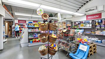 Een winkel met gangpaden met de aanduiding 'SNACKS' en 'HYGIEN', manden met producten, en een blauwe plastic stoel.