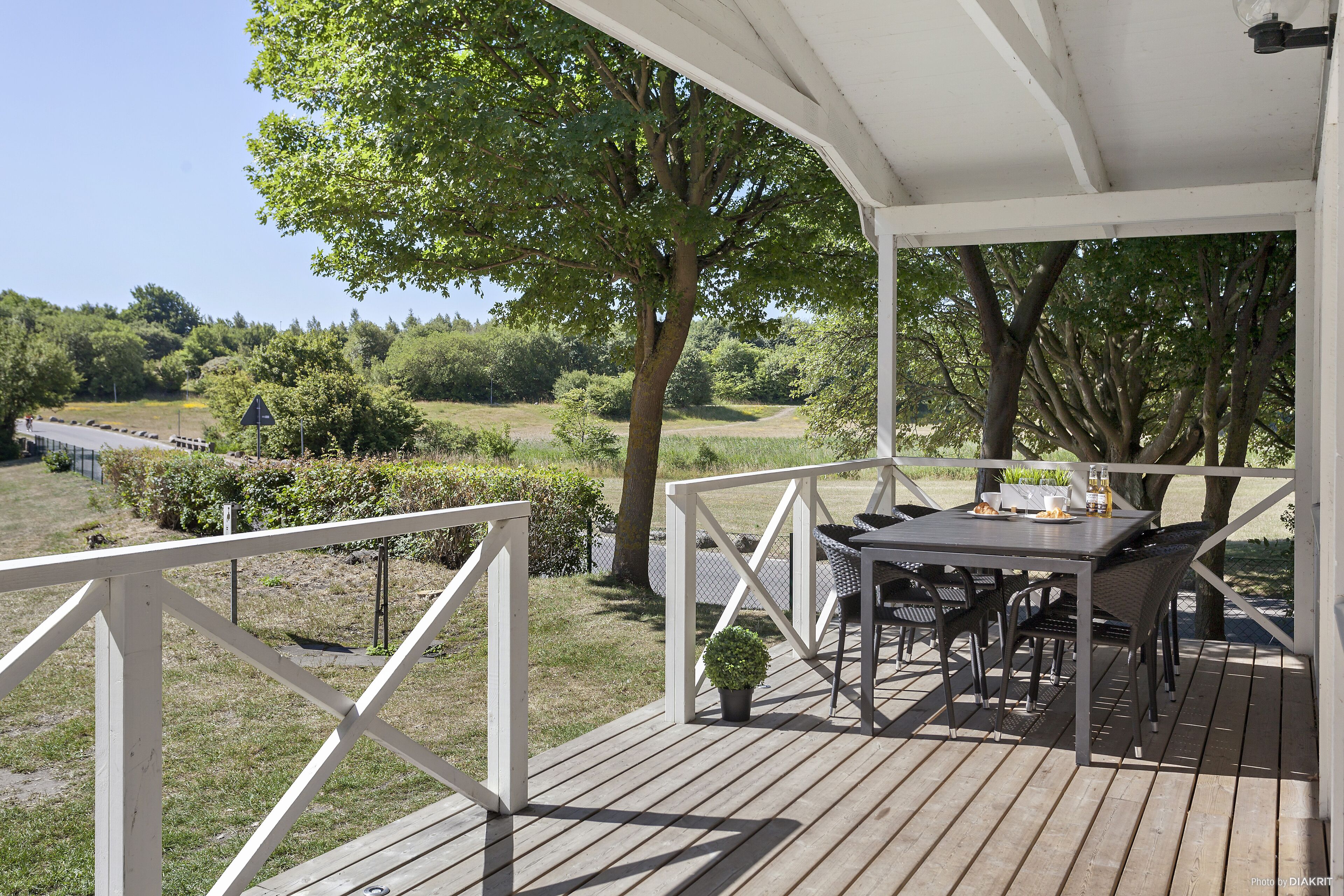 Une terrasse en bois avec une table et des chaises, entourée de rampes blanches et d’arbres.