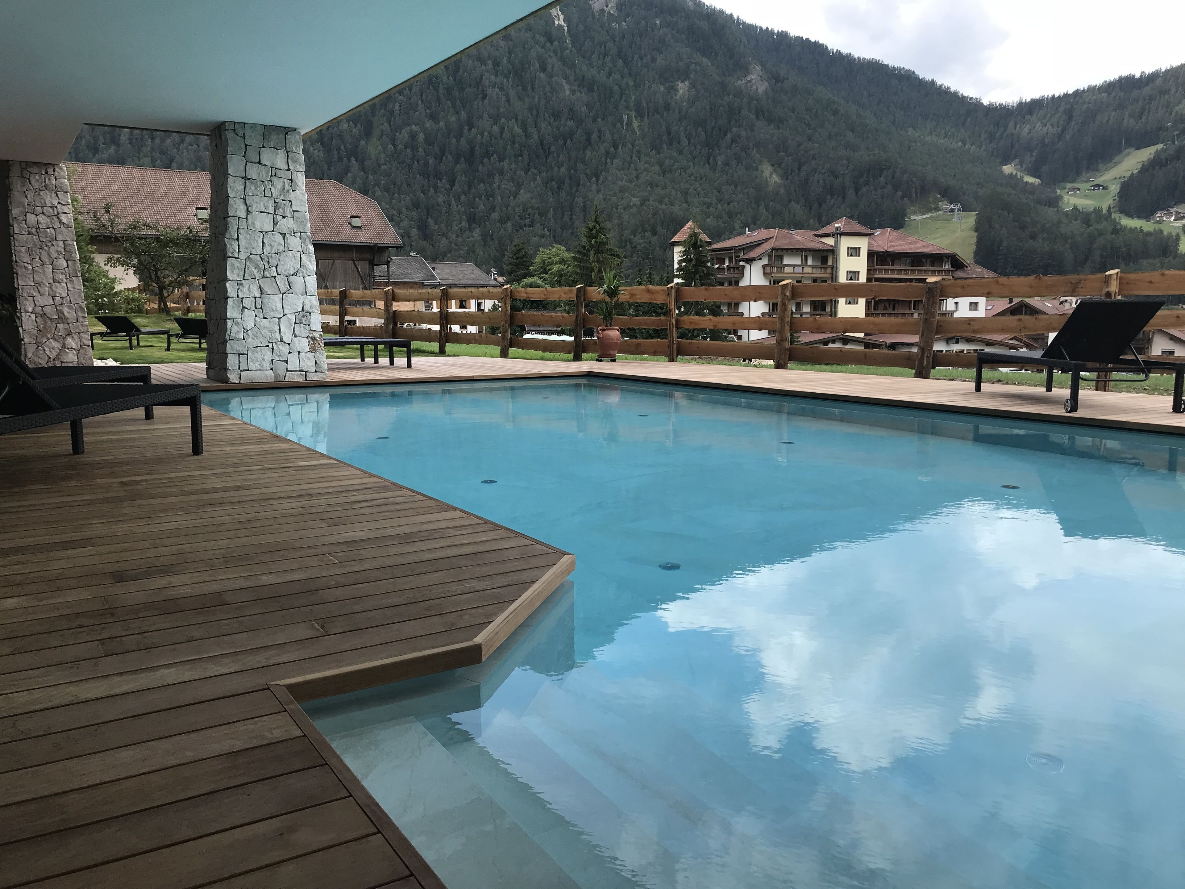 A pool area with wooden deck and lounge chairs, surrounded by stone pillars and a wooden fence.